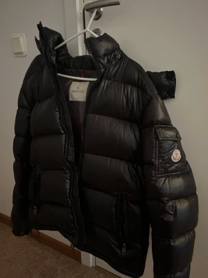 Svart pufferjacka från Moncler - Snygg svart pufferjacka från Moncler med glansig finish, huva och dragkedjor på fickorna. Klassisk Moncler-logga på ärmen och breda kanaler för extra värme. Perfekt för kalla vinterdagar och riktigt trendig stil.