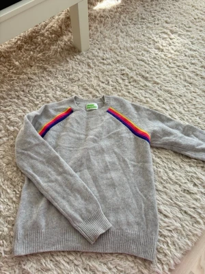 from future tröja - Säljer en grå stickad tröja från From Future med färgglada regnbågsränder på axlarna. Tröjan har rund halsringning, långa ärmar och ribbstickade muddar. Perfekt för dig som gillar färg och vill ha något unikt i garderoben.