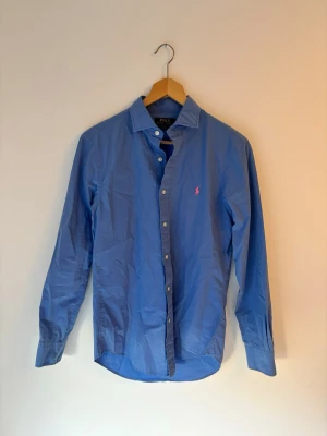 Blå skjorta från Polo Ralph Lauren - Klassisk blå skjorta från Polo Ralph Lauren i slim fit-modell. Skjortan har långa ärmar, knappar framtill och den ikoniska rosa logotypen broderad på bröstet. Tillverkad i mjuk bomull som känns skön mot huden. Skjortan är sprillans ny och har aldrig använts