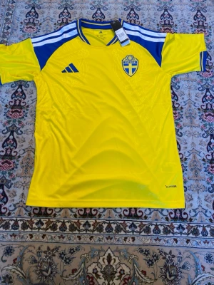 Svensk landslagströja Adidas gul/blå - Säljer en  svensk landslagströja  i storlek XL. Tröjan är gul med blå detaljer och har Sveriges fotbollsemblem på bröstet. Materialet är lätt och andas, perfekt för sport och träning. Klassisk rund hals och Climalite-teknologi.