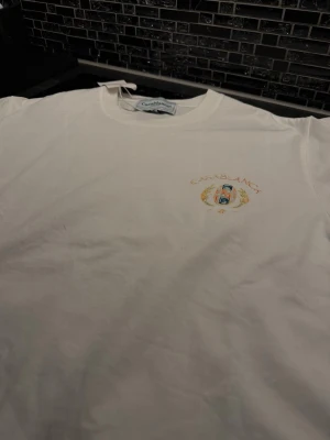 Casablanca t-shirt - Säljer en vit t-shirt från Casablanca med färgglad 'Tennis Club'-print på bröstet och ryggen. Perfekt för dig som gilla clean stil till sommaren som kommer.