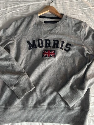 Grå sweatshirt från Morris - Grå sweatshirt från Morris med broderad text och brittisk flagga på bröstet. Tröjan har rund halsringning, ribbade muddar och nederkant samt en patch på ärmen. Perfekt för en avslappnad och stilren look.