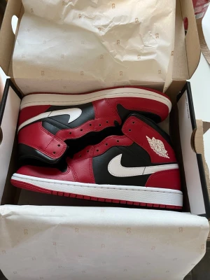 Air Jordan 1 High röd/svart/vit - Säljer ett par Air Jordan 1 High sneakers från Nike i klassisk röd, svart och vit färgkombination. Skorna har högt skaft, vit swoosh och ikonisk Air Jordan-logga på sidan. Tillverkade i läder med snygga detaljer och levereras i originalkartong. KAN LÄTT GÅ NER I PRIS!