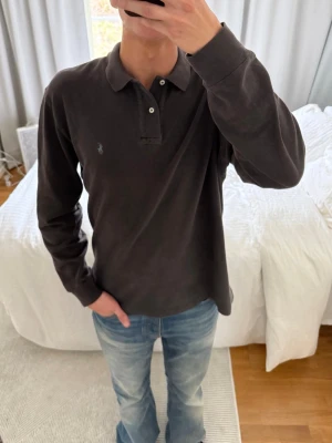 Mörkgrå långärmad piké från Polo Ralph Lauren - Säljer en mörkgrå långärmad pikétröja från Polo Ralph Lauren med klassisk krage och två knappar framtill. Diskret logga broderad på bröstet. Perfekt för dig som gillar stilrena och tidlösa plagg.