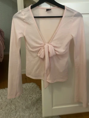 Ljusrosa topp med knyt - Säljer en ljusrosa topp med knyt framtill. Toppen har v-ringning och den är öppen i magen med långa ärmar, perfekt för sommaren. Materialet känns mjukt och lätt, vilket gör den bekväm att bära. Passar dig som gillar stilrena och feminina plagg.
