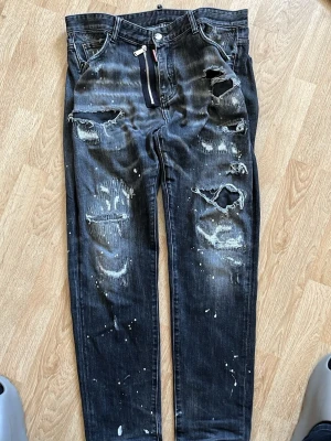 Svarta distressed jeans från Dsquared2 - Coola svarta jeans från Dsquared2 med slitna detaljer, stora hål och vita färgstänk. Klassisk femficksmodell med silverfärgad dragkedja och röd logotyp-tag. Jeansen har en rak passform och är tillverkade i denim.