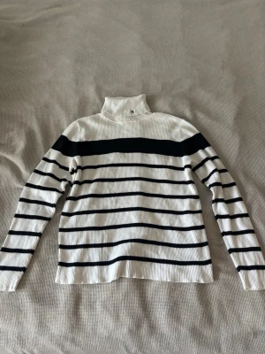 Randig polotröja från Ralph Lauren - Säljer en vit ribbstickad polotröja från Ralph Lauren med svarta horisontella ränder. Tröjan har hög krage med diskret RLL-brodyr och långa ärmar. Perfekt för dig som gillar klassisk och stilren design.