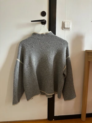 Zara tröja  - En grå stickad tröja med vita detaljer längs kanterna och sömmarna. Tröjan har en hög hals och långa ärmar, perfekt för att hålla sig varm under kyliga dagar. Passformen är loose och materialet känns mjukt och bekvämt.