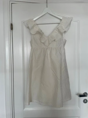 Vit klänning - Säljer en vit klänning med v-ringning och stora volanger längs axlarna. Klänningen är ärmlös och har en lätt strukturerad yta. Ny med lapp kvar, endast testad!