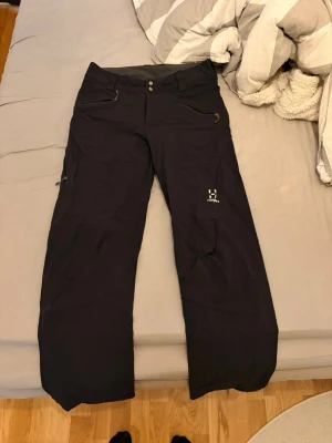 Svarta täckbyxor från Haglöfs - Täckbyxor från Haglöfs i svart, perfekta för kalla dagar. Byxorna har två knappar i midjan, dragkedja, fickor med dragkedja och logga på benet. Materialet är slitstarkt och designen är rak med förstärkta knän. Grymma för utomhusaktiviteter och vinterväder.