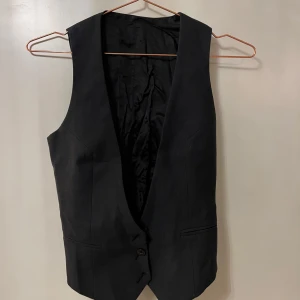 Svart väst från J.Lindeberg Denim - Klassisk svart väst från J.Lindeberg Denim med djup v-ringning och knäppning framtill. Västen har två fickor fram och är helfodrad med slät insida. Perfekt att styla över en skjorta eller t-shirt.