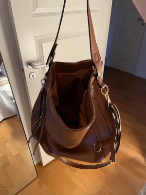 Brun axelväska i skinnimitation - Snygg brun axelväska i skinnimitation med justerbart axelband och guldfärgade metalldetaljer. Väskan har en rymlig insida, perfekt för dig som vill ha plats för allt du behöver under dagen.