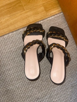 Svarta pumps med flätad rem från Ellos - Snygga svarta pumps från Ellos med två breda flätade remmar i svart och guld. Skorna har fyrkantig tå och låg klack, vilket ger en trendig look. Materialet ser ut att vara konstläder och insidan är ljusbeige. Perfekta för dig som vill sticka ut.
