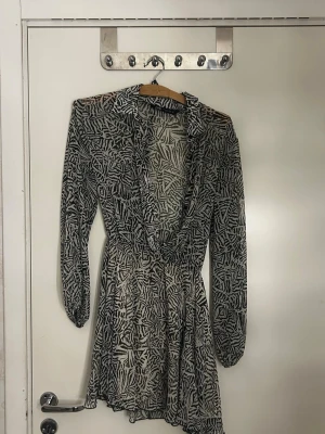 Svartvit mönstrad omlottklänning - Kort omlottklänning med svartvitt abstrakt mönster. Klänningen har långa ärmar med resår vid handlederna och en djup v-ringning. Tillverkad i ett tunt, lätt och något transparent material som ger ett luftigt intryck. Perfekt för dig som gillar trendiga och unika plagg.