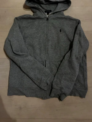 Grå hoodie från Polo Ralph Lauren - Snygg grå zip-hoodie från Polo Ralph Lauren med klassisk svart logga på bröstet. Hoodien har dragkedja, två fickor framtill och mjukt material som känns skönt mot huden. Perfekt för en avslappnad och stilren look. Storlek Xl men passar mer L 