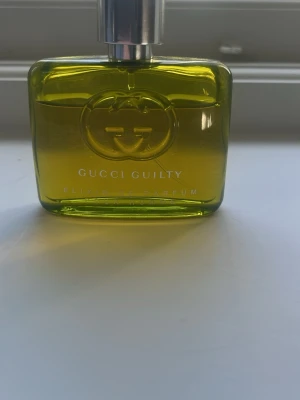 Gucci Guilty Elixir Eau de Parfum - Gucci Guilty Elixir Eau de Parfum i en stilren, rektangulär flaska med metallisk silverfärgad kork. Flaskan är transparent med en intensiv gulgrön nyans och har Guccis ikoniska logotyp i relief på framsidan. Perfekt för dig som vill ha en lyxig och unik doft.
