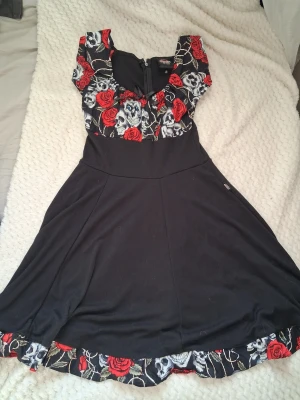 ~Rockabilly klänning skull & roses-/goth retro~ - Rockabilly klänning med färgerna svart,vit och röd  har döskallar på sig.Perfekt för dig som gillar alternativ, goth eller edgy stil 🌹💀  Storlek: XS, står 10-12 år men passar mig med storlek XS-S.  Skick: Fint använt skick  Passform: Markerad midja och utsvängd kjol (väldigt smickrande!)  Dragkedja baktill!  Passar både till vardag och fest – lätt att styla med boots eller sneakers 👢 