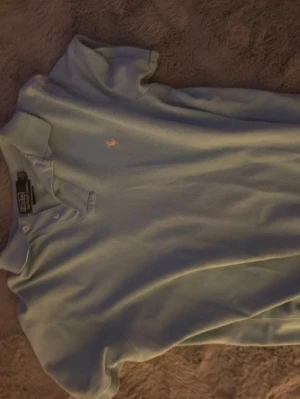 Ljusblå pikétröja från Polo Ralph Lauren - Klassisk ljusblå pikétröja från Polo Ralph Lauren med vit broderad logga på bröstet. Tröjan har krage, knappar vid halsen och korta ärmar. Perfekt för en avslappnad men ändå stilren look.