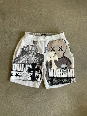 Gråa Ken2 shorts med tryck - Unika gråa shorts från Ken2 med elastisk midja och stora grafiska tryck på båda benen. Motiven inkluderar dödskalle, kors, stjärnor och texten 'WORLDWIDE'. Perfekta för dig som gillar streetwear och statement-plagg.