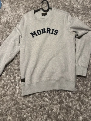 Grå sweatshirt från Morris - Snygg grå sweatshirt från Morris med mörkblå broderad logotyp på bröstet. Tröjan har rund halsringning, ribbade muddar och är tillverkad i mjukt bomullsmaterial. Perfekt för en avslappnad och cool look.