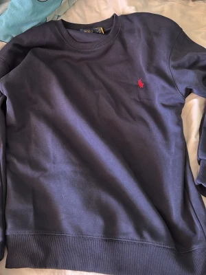 Mörkblå sweatshirt från Polo Ralph Lauren - Mörkblå sweatshirt med rund hals från Polo Ralph Lauren. Tröjan har en klassisk passform och är dekorerad med det ikoniska röda broderade logot på bröstet. Perfekt för en avslappnad och stilren look.