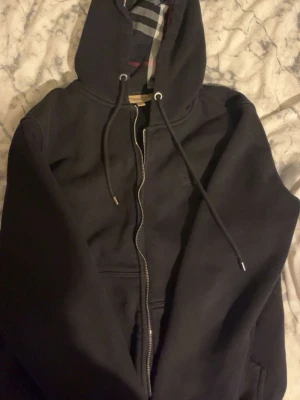 Svart Burberry zip - Svart hoodie från Burberry med klassiskt rutigt foder i huvan och dragkedja framtill. Modellen har snören i huvan och ribbade muddar. Materialet är mjukt och bekvämt, perfekt för en avslappnad stil. Ikoniskt Burberry-mönster på insidan av huvan.