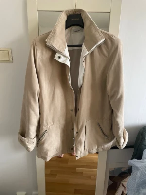 Beige mockajacka från Cappuccini – stilren vår/höstjacka - Snygg och tidlös jacka från Cappuccini i mjuk mockaimitation (eller tunn mocka-look). Klassisk modell med hög krage, dragkedja och tryckknappar framtill. Längs ner på insidan av jackan liten färg ,som ni kan se på sista bilden 