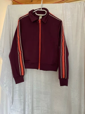 Vinrött träningsset med ränder - Snyggt vinrött träningsset med zip-jacka och matchande byxor. Jackan har orange dragkedja och ränder i orange och vitt längs ärmarna. Byxorna har raka ben och orange snörning i midjan. Perfekt för sport eller chill. Materialet är mjukt och stretchigt.