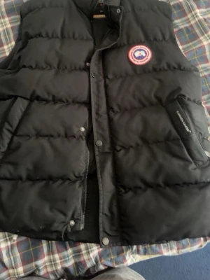 Svart dunväst från Canada Goose - Säljer en svart dunväst från Canada Goose med klassisk logotyp på bröstet. Västen har dragkedja och knappar framtill samt två snedställda fickor med Canada Goose-brodyr. Perfekt för lager-på-lager och håller dig varm under kyliga dagar. Pris kan diskuteras vid snabb affär!