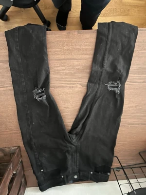Svarta slitna jeans med hål - Svarta jeans med skinny fit och slitna detaljer på båda knäna. Jeansen har klassisk femficksdesign och dragkedjegylf. Perfekta för en edgy streetstil.