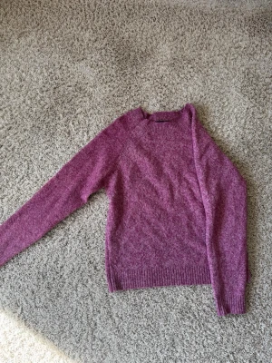 Lila stickad tröja med rund hals - Mysig lila stickad tröja med rund hals och långa ärmar. Tröjan har ribbstickade muddar vid ärmslut och nederkant. Perfekt för att hålla värmen under kyliga dagar och passar till både jeans och kjol. Färg finns ej att köpa i butik längre.