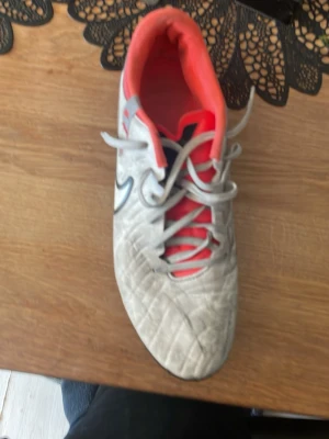 Nike vita fotbollsskor med dobbar - Säljer ett par vita Nike fotbollsskor med svarta och röda detaljer. Skorna har klassisk snörning, dobbar för bra grepp på gräsplan och en vadderad, röd insida för extra komfort. Perfekta för dig som vill ha stilrena och funktionella skor på planen.