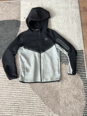 Nike Tech Fleece hoodie svart/grå - Snygg Nike Tech Fleece hoodie i svart och ljusgrått med dragkedja framtill och huva. Hoodien har en modern design med kontrastpaneler, två sidofickor och Nike-logga på bröstet. Perfekt för en sportig och avslappnad stil. Tillverkad i mjukt och stretchigt material.