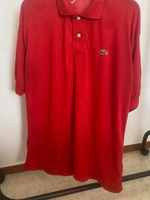 Röd pikéskjorta från Lacoste - Säljer en klassisk röd pikéskjorta från Lacoste med den ikoniska krokodilloggan på bröstet. Skjortan har krage, knappar framtill och korta ärmar. Perfekt för dig som gillar en sportig och stilren look.