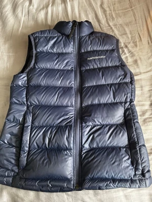 Blå dunväst från Peak Performance - Snygg blå dunväst från Peak Performance med hög krage och dragkedja framtill. Västen har ett quiltat mönster och två sidofickor. Perfekt för lager-på-lager och håller dig varm under kyliga dagar. Logotypen är diskret broderad på bröstet.                                                                             Ny pris ca 1800kr 