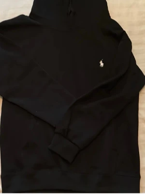 Svart hoodie från Polo Ralph Lauren - Svart hoodie från Polo Ralph Lauren med klassisk huva och dragsko. Diskret vit logga broderad på bröstet. Mjuk bomullskänsla och ribbade muddar vid ärmslut och nederkant. Perfekt för en chill och stilren look.