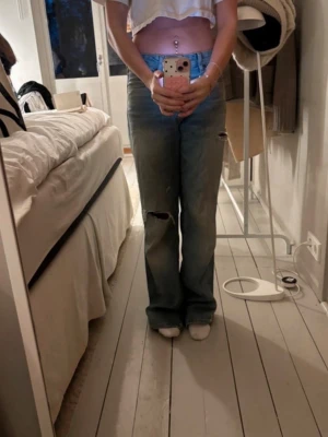 Blå slitna jeans med raka ben - Säljer ett par ljusblå jeans med raka ben och slitningar på knäet. Jeansen har klassisk femficksdesign. Perfekt för dig som gillar en avslappnad och trendig look.