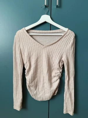 Tröja - Trendig beige ribbad topp med v-ringning och långa ärmar. Snygg rynkdetalj på sidan ger en cool twist. Perfekt att styla med jeans eller kjol för en avslappnad look.
