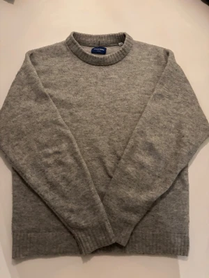 Grå stickad tröja från Jack & Jones - Klassisk grå stickad tröja från Jack & Jones med rund halsringning och ribbade muddar vid ärmslut och nederkant. Perfekt för lager-på-lager och enkel att matcha med andra plagg. Mjuk och skön känsla.