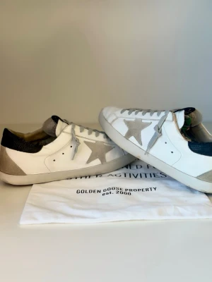 Golden Goose vita sneakers med stjärna - Snygga vita sneakers från Golden Goose med grå mockadetaljer och den ikoniska stjärnan på sidan. Skorna har svarta hälpartier och grå snörning. Tillverkade i läder och mocka för en cool och avslappnad look.