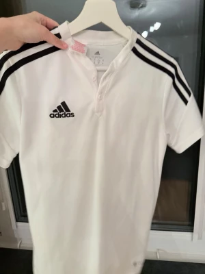 Vit Adidas sporttröja med svarta ränder - Snygg vit sporttröja från Adidas med klassiska svarta ränder på axlarna och Adidas-logga på bröstet. Tröjan har korta ärmar och knappar vid halsen. Perfekt för träning eller sportiga outfits.