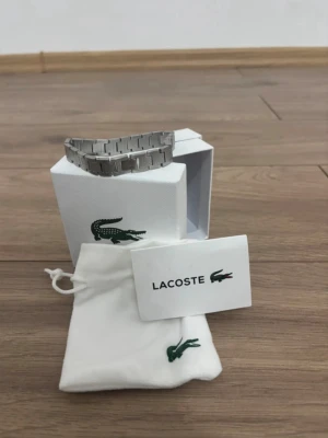 Armband från Lacoste - Snyggt armband i silverfärgad metall från Lacoste med klassisk krokodil-logga ingraverad på länkarna. Armbandet har en stilren och modern design med rektangulära länkar och kommer i originalförpackning med tygpåse och certifikat. Kontakta gjärna vid frågor😘