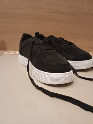 Svarta sneakers med vit sula - Stilrena svarta sneakers med tjock vit sula och klassisk låg profil. Ovandelen är i mockaliknande material och skorna har svarta skosnören. Perfekta för dig som gillar en clean och modern look.