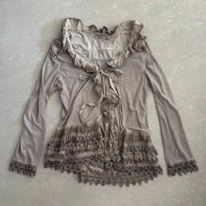 Beige volangkofta med spetsdetaljer - Kofta med volanger och dekorativa spetsdetaljer längs kanter och ärmslut. Koftan har knytband framtill och långa ärmar!❤️