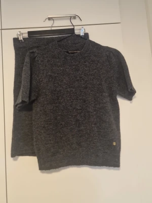 Mörkgrått stickat set från Pernilla Wahlgren  - Snyggt tvådelat set från Pernilla Wahlgren i mörkgrå stickad kvalitet. Setet består av en kortärmad topp med rund hals och en matchande kjol med resår i midjan. Båda delarna har en liten guldfärgad logodetalj. Perfekt för en trendig och bekväm look.