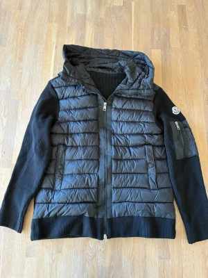 Moncler cardigan - Snygg svart cardigan från Moncler med vadderad framsida och huva i. Ärmar och rygg i ribbstickad bomull, dragkedja framtill och två fickor med knapp. Moncler-logga på ärmen och extra ficka med dragkedja på vänster arm.