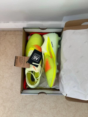 Nike Phantom 6 Low Elite AG - Snygga Nike Phantom GX Elite fotbollsskor i neon-gult med orange och svarta detaljer. Skorna har AG-yttersula för konstgräs, snörning och textilöverdel med struktur för bättre bollkontroll. Perfekta för dig som vill sticka ut på planen.