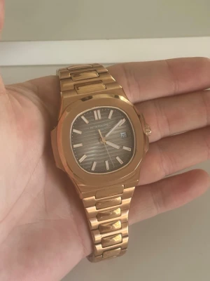 Patek Philippe Nautilus  - Exklusiv Patek Philippe Nautilus klocka i guld med länkarmband och mörk urtavla med horisontella linjer. Klockan har datumvisning och tydliga indexmarkeringar. En riktigt lyxig accessoar som sticker ut och passar till alla tillfällen. Extra länkar följes med!