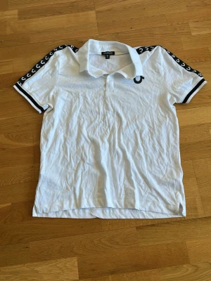 Vit pikétröja från C.P. Company - Vit pikétröja från C.P. Company med svarta detaljer och logga på bröstet. Svarta ränder på ärmsluten och dekorativa band med logotyp längs axlarna. Klassisk krage och korta ärmar. Perfekt för en sportig och stilren look.