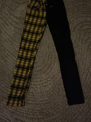 Gula och svarta rutig jeans Banned - Unika jeans från Banned Alternative med ena benet i gul och svart rutig design och andra benet helsvart. Snygga detaljer med ringar och hög midja. Perfekta för dig som vill sticka ut med din stil.